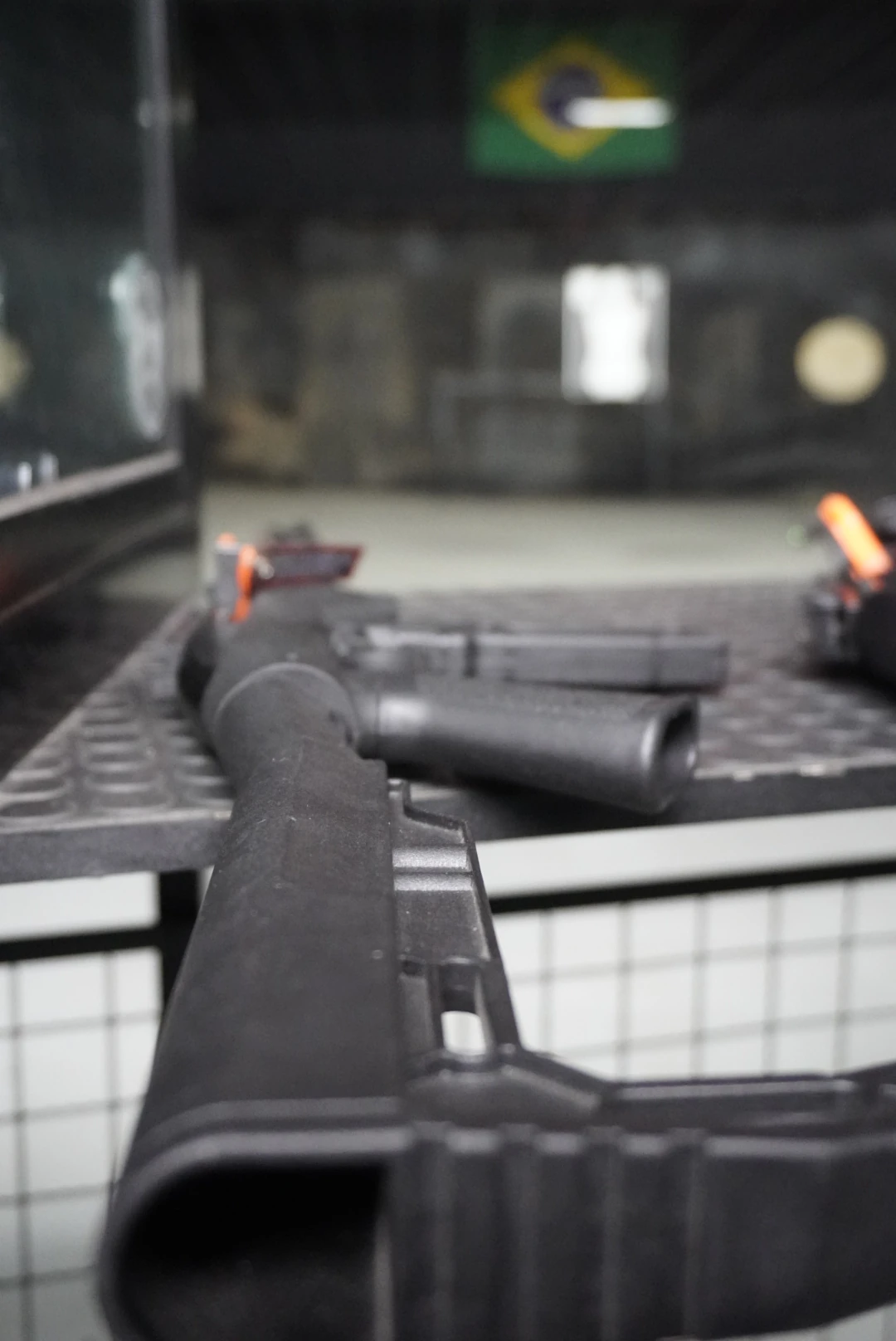 Pistolas e carabina apoiadas na bancada da baia do Clube de Tiro Sumrak 408 com bandeiras de segurança laranja
