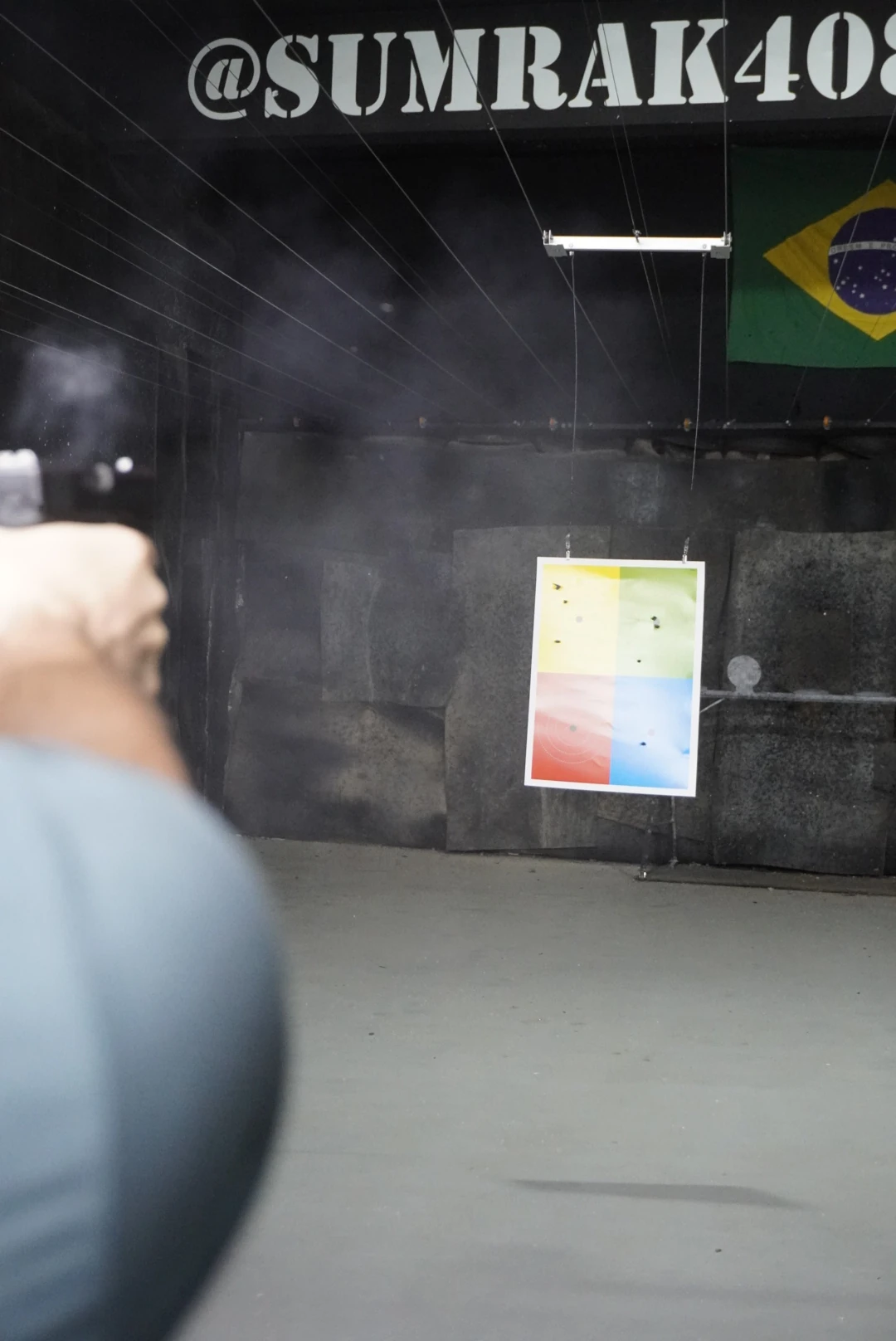 Atirador em posição na raia do Clube de Tiro Sumrak 408 mirando alvo colorido suspenso, com fumaça de disparo e bandeira do Brasil ao fundo