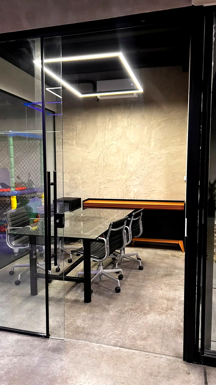 Sala de coworking envidraçada da Sumrak 408 com luminária linear no teto, mesa de vidro, cadeiras executivas e vista para o Espaço Kids