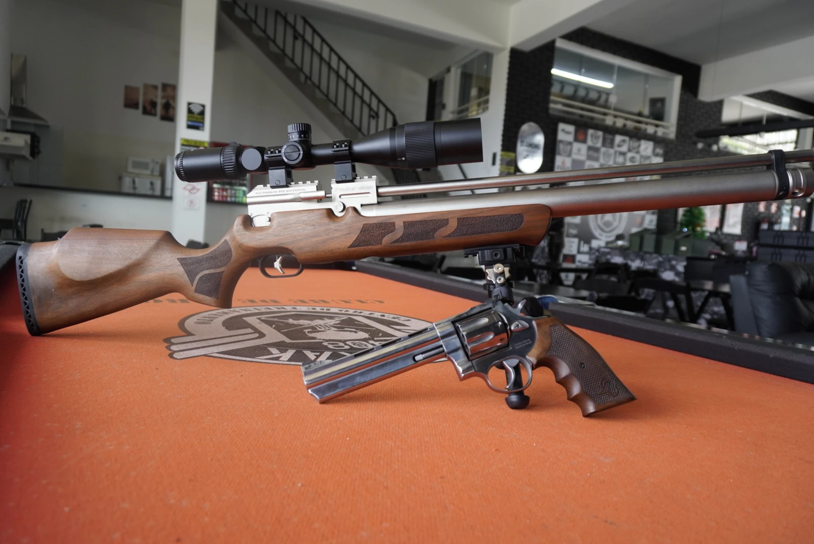 Carabina PCP com luneta e revólver de aço inox expostos sobre mesa laranja na loja do Clube de Tiro Sumrak 408