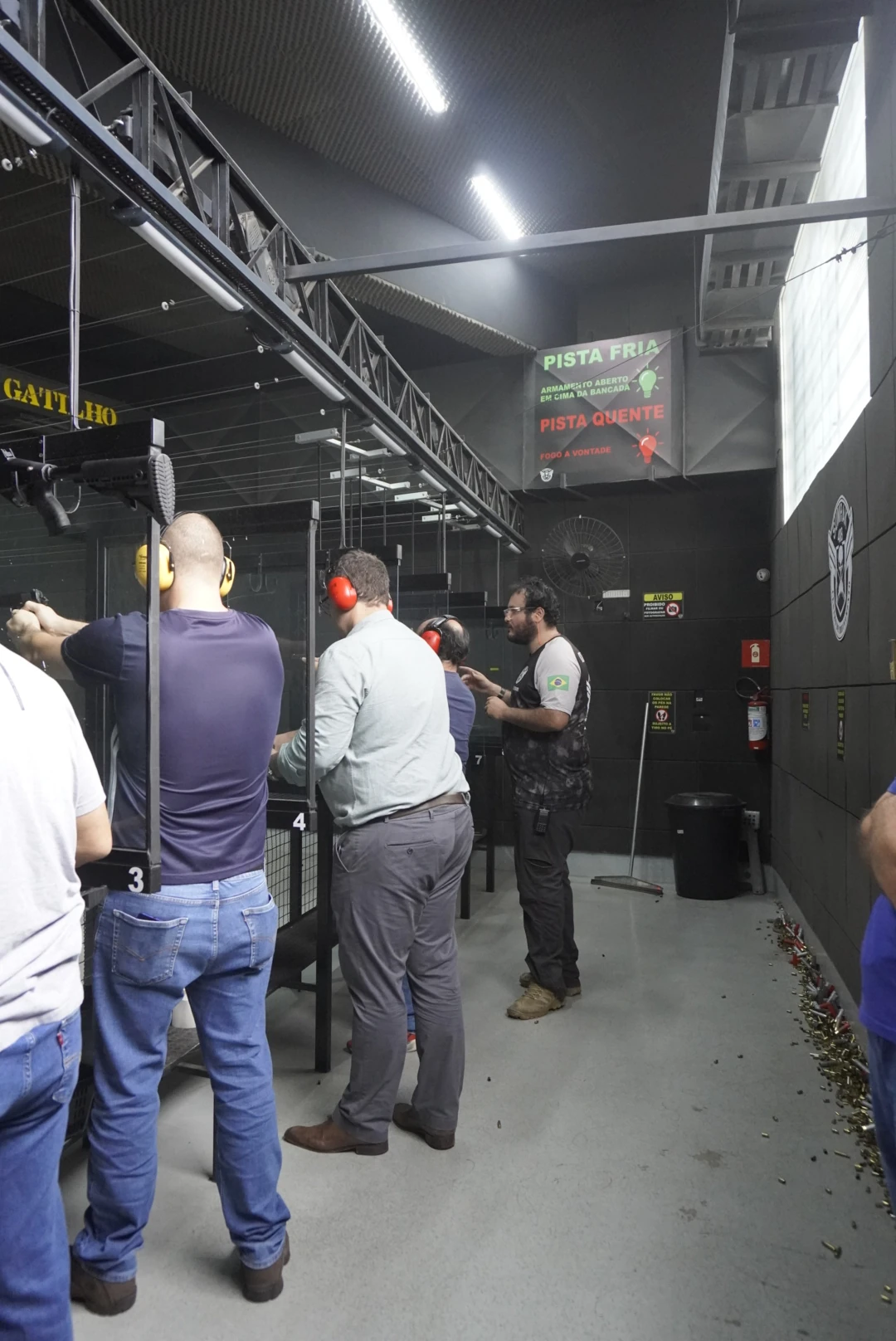 Instrutor acompanhando atiradores nas baias do Clube de Tiro Sumrak 408, com placas de pista fria e pista quente ao fundo