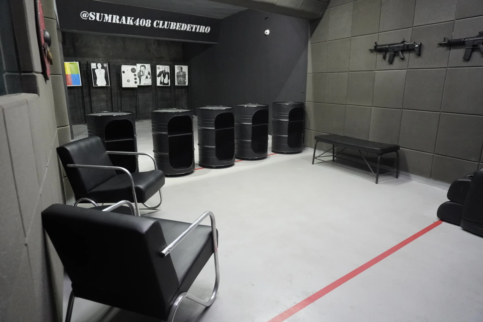 Área de espera do Clube de Tiro Sumrak 408 com poltronas, tambores metálicos, alvos expostos na parede e piso demarcado em vermelho